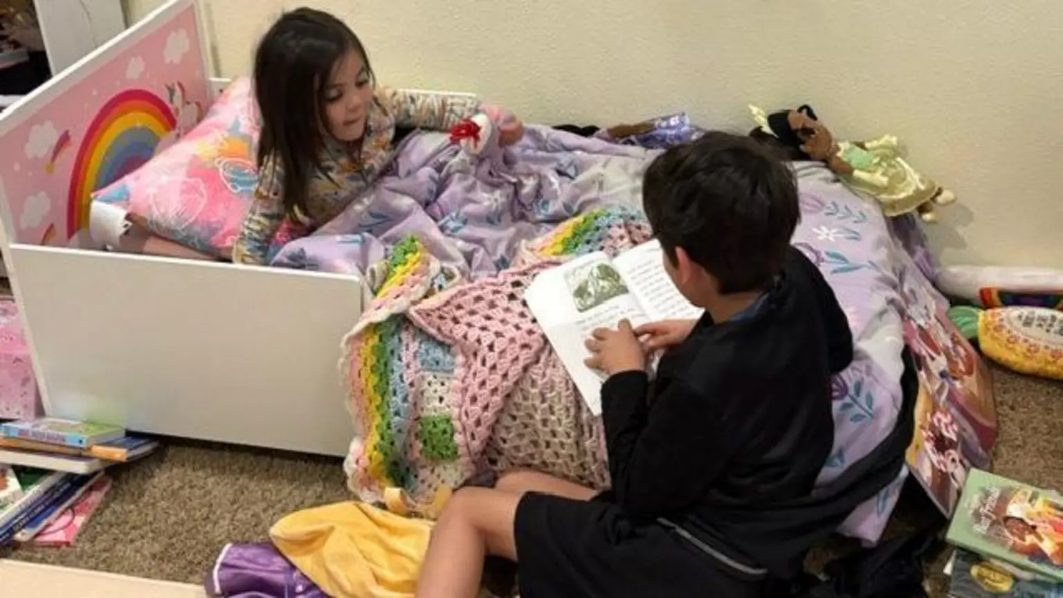 A young boy clad in black shorts and a black top sits on the ground, holding a book on the edge of his sister's bed as he reads it to her; she sits under a purple blanket, a rainbow visible on the headboard