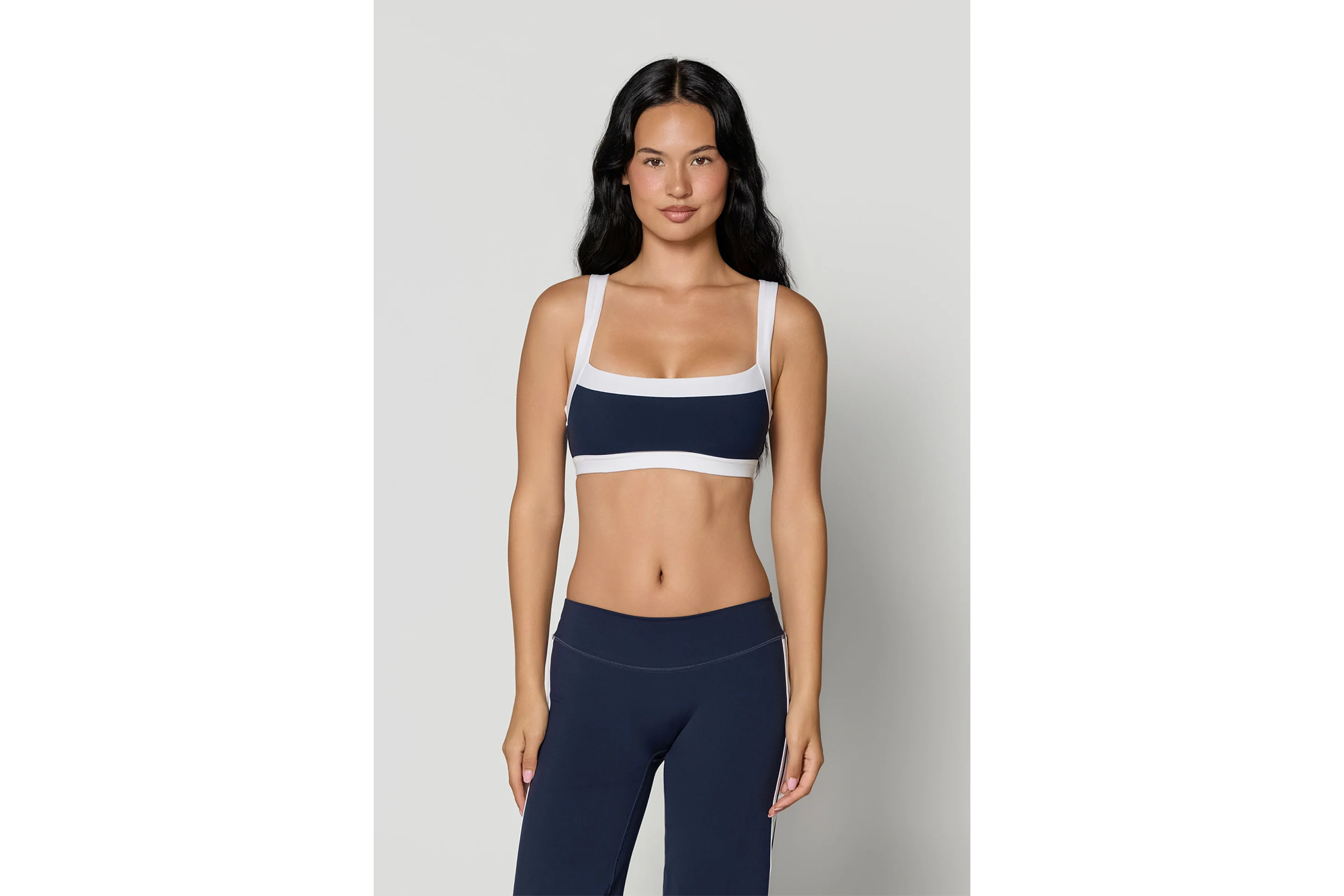 A woman with long dark hair stands facing forward, wearing a dark blue sports bra with white straps and trim, and dark blue leggings with a white stripe down the side.