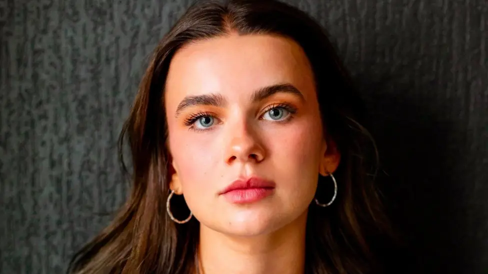 Tabitha Smyth has long brown hair looks to the camera as she wears hooped gold earrings. She has green eyes and stands in front of a black-grey wall.