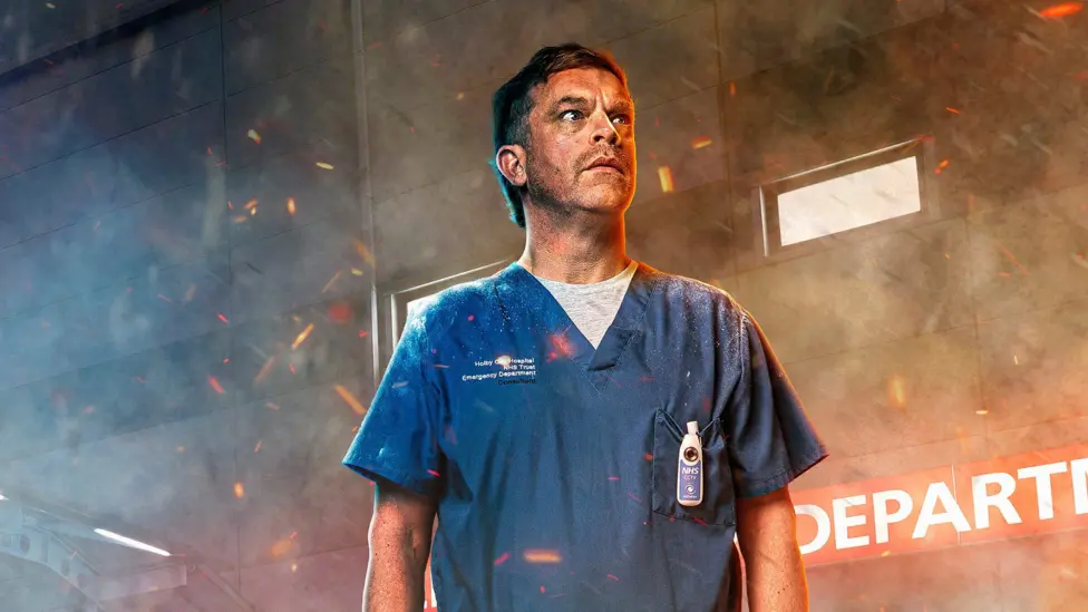 Promotional image of William Beck in character as Dylan Keogh. He is photographed from the waist up and is wearing blue medical scrubs. The set of the emergency department can be seen behind him. His face is covered in s