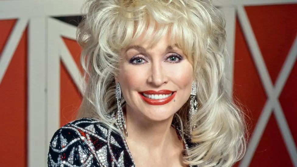 A head and shoulders shot. Dolly's blonde hair stands up in big waves and falls onto her shoulders. She wears a sliver and black diamante V neck dress with red accent colours. Long diamante earrings hang down.
