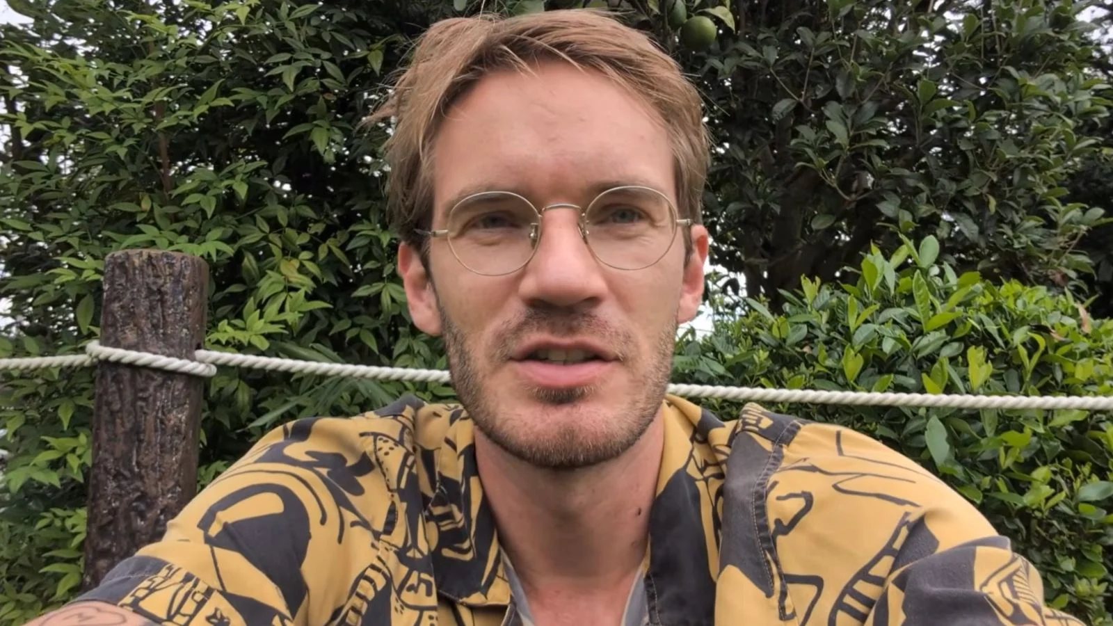 PewDiePie in glasses and gold shirt talking to camera near trees