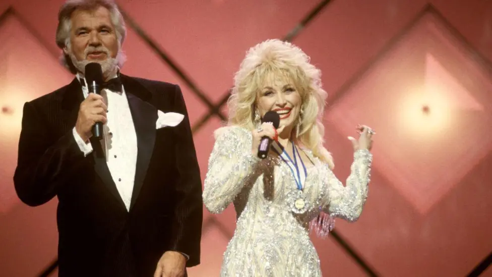 Kenny Rogers, wearing a black and white tuxedo, and Dolly Parton, wearing a bejewelled white and cream long sleeved dress, performing on stage with a pink background.