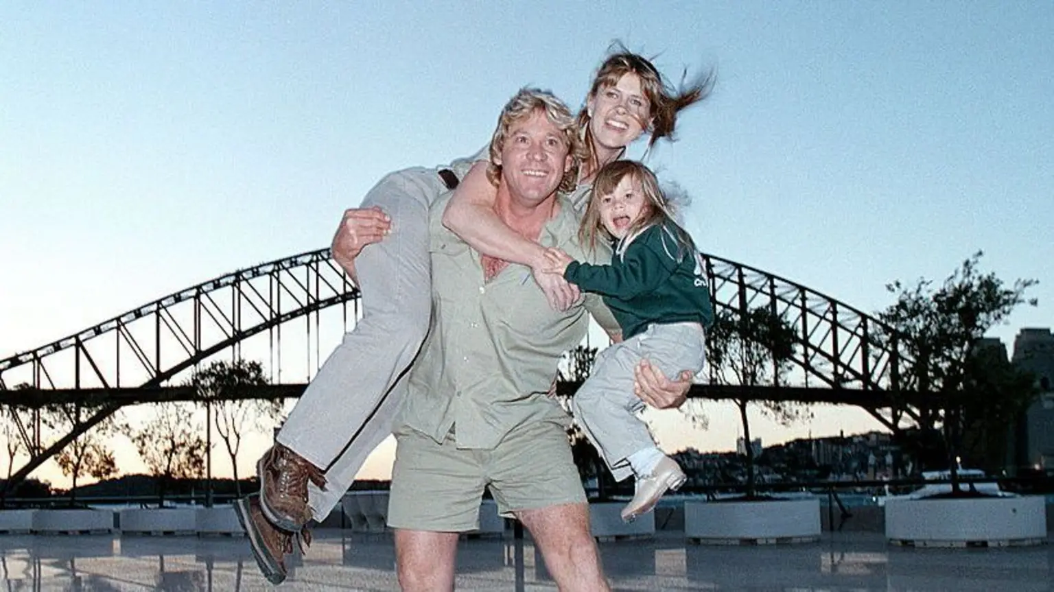 A man wearing khaki shirt and shorts holding a young child with one arm and an adult woman with his other arm with the Sydney Harbour Bridge in the background