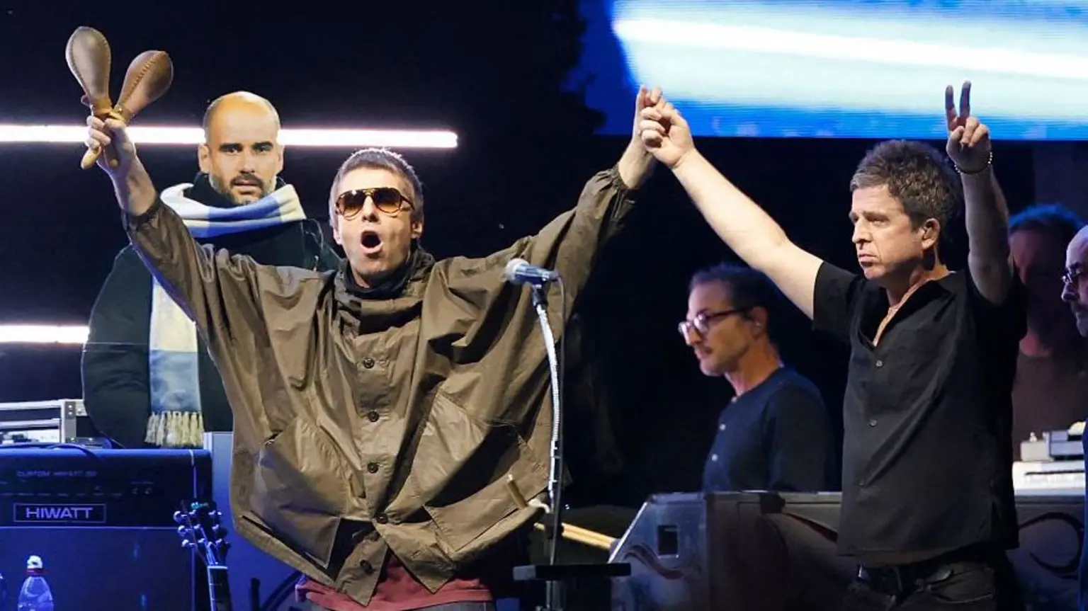 Noel and Liam Gallagher at one of the Oasis reunion gigs. They are holding their arms in the air and touching hands in the middle. Liam is holding maracas in his other hand while Noel is doing a peace sign with his finge
