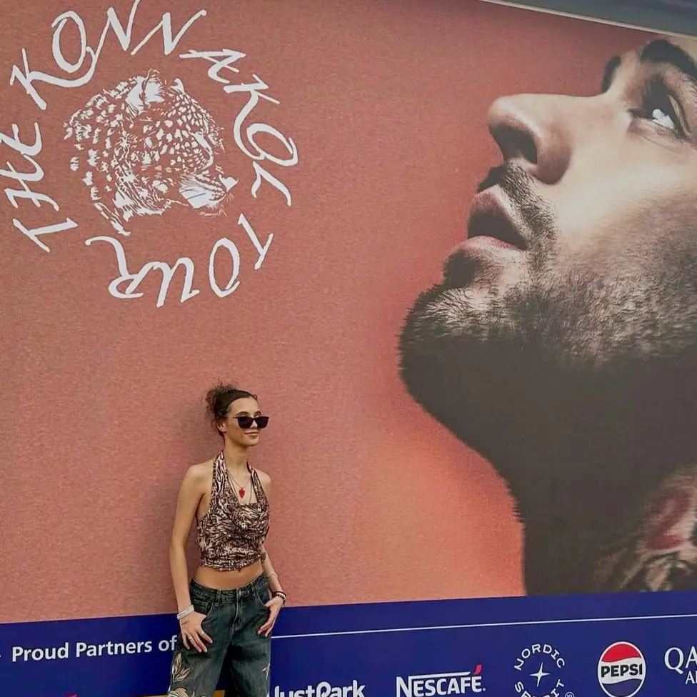 Aurélia Collins stands in front of a billboard of Zayn Malik promoting his tour. She is wearing a halter-neck top, jeans and sunglasses.