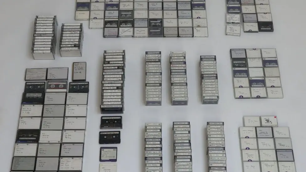 Dozens of tapes are grouped together and laid out on a white surface. There is small hand written notes on each tape.