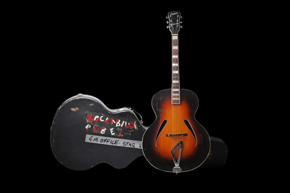 An acoustic guitar with a sunburst pattern, standing next to a hard, black guitar case with the words Rockabilly Rebel written on it in red and white capital letters. The case and guitar are against a black background.