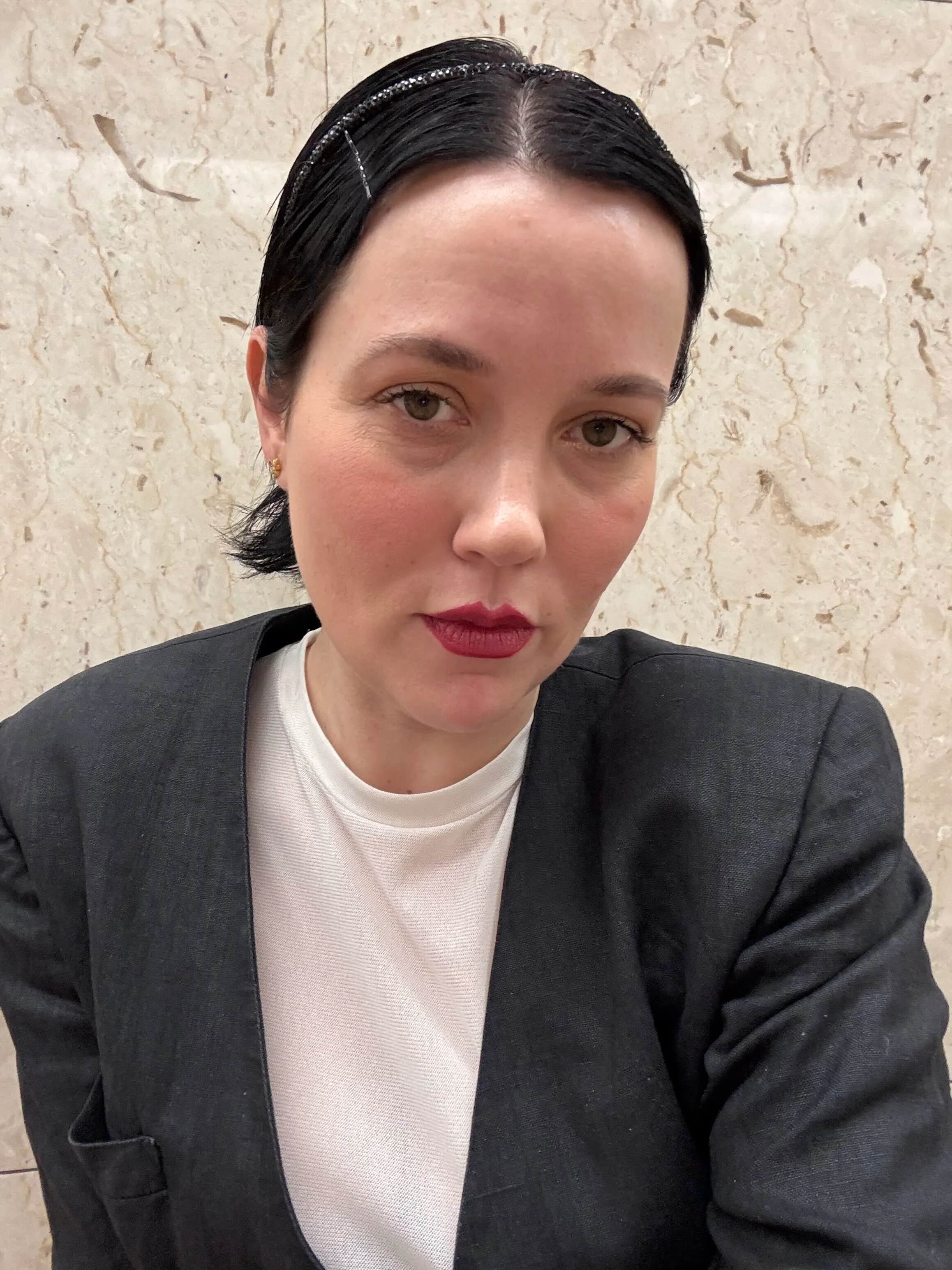 A woman with dark hair, a black jacket, and a white t-shirt looks directly at the camera.