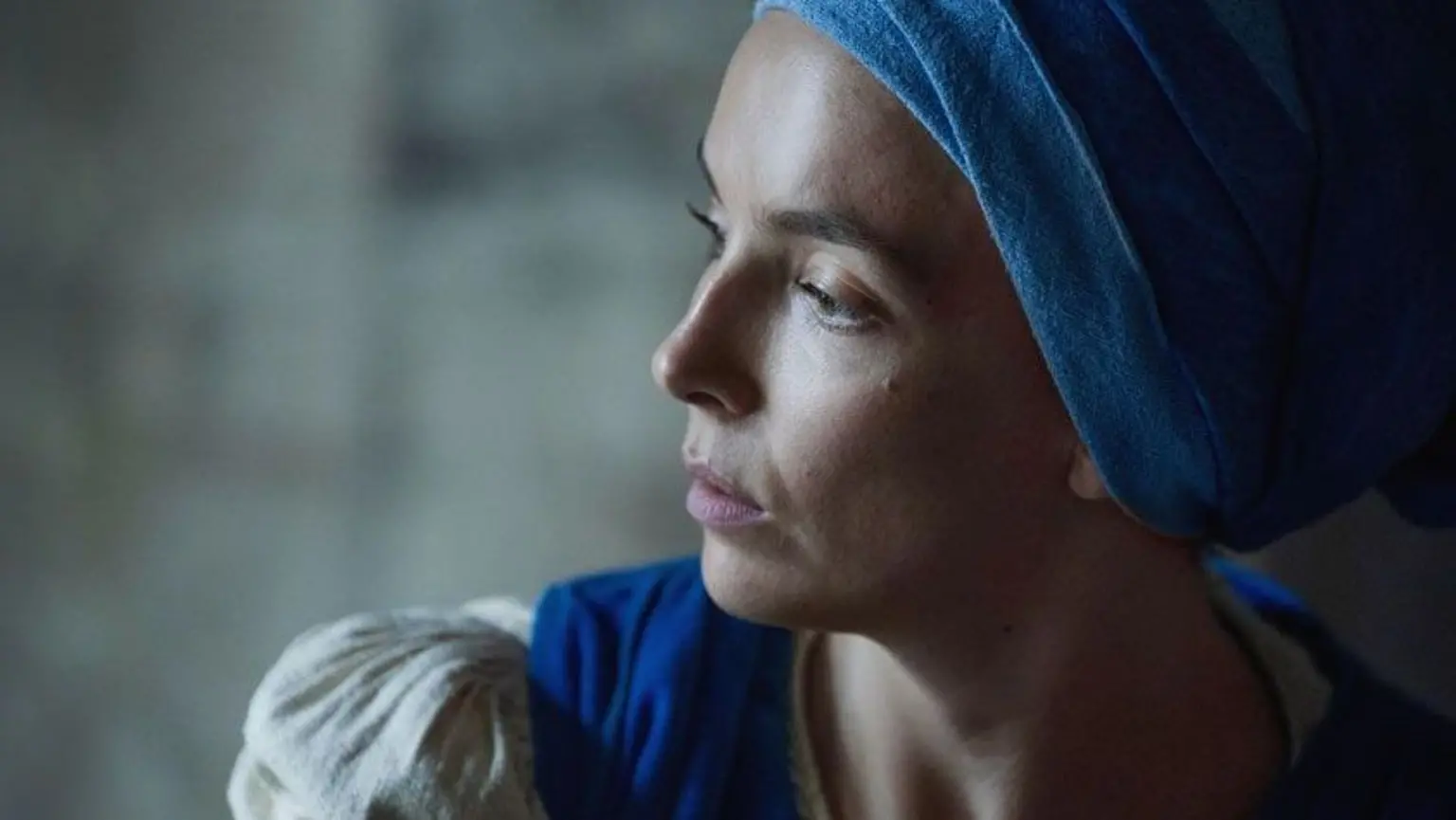 Side view of Jodie Comer. She is wearing a blue head wrap, a beige dress with a blue apron.