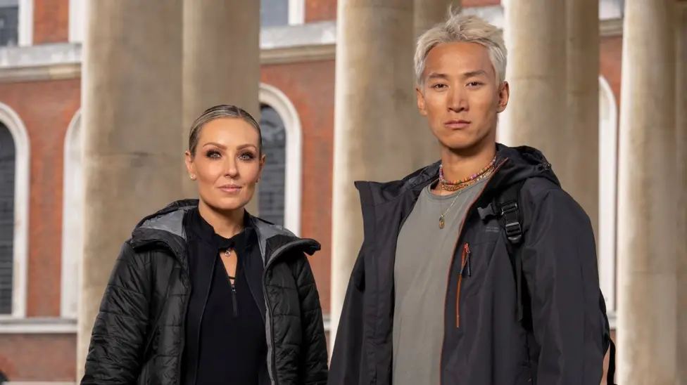 Carlos Gu and Amy Dowden pictured side by side in a promotional photo for the Channel 4 show, Celebrity Hunted. Carlos is wearing a grey dark coat, a light grey t-shirt and several necklaces. Amy is wearing a black top,