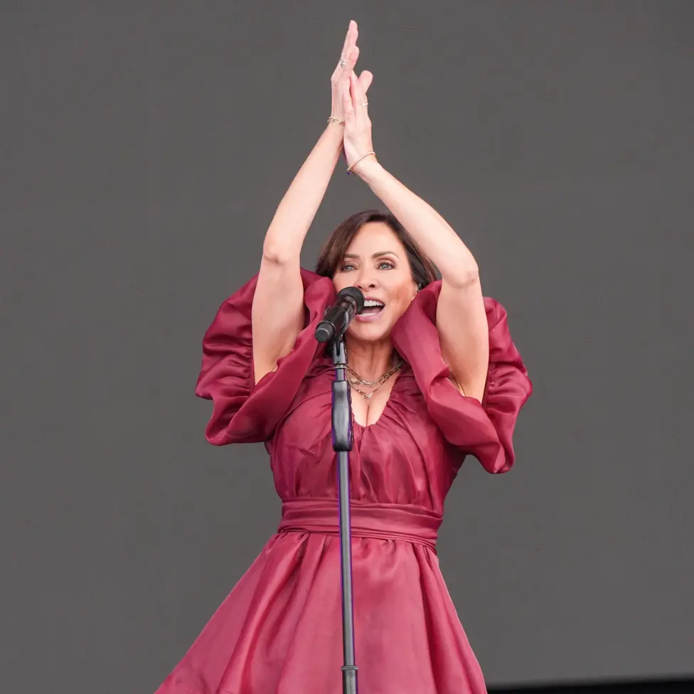 Natalie Imbruglia performs on stage in a cherry red ballgown, with her hands raised above her head as she encourages the audience to clap