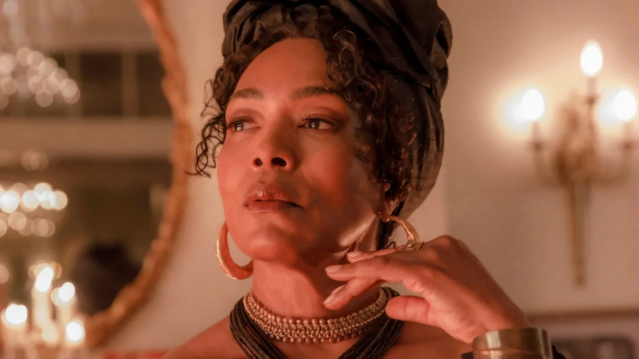A woman in a dark head wrap, large gold hoop earrings, and layered beaded necklaces touches her chin, softly lit by candlelight.