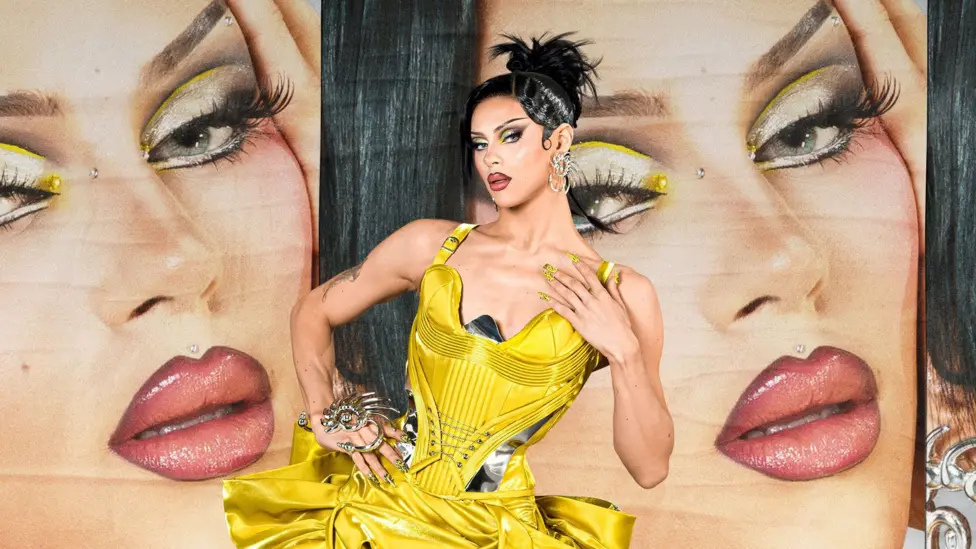 Flesh wears a yellow strappy dress and has a dark brunette wig on done in an up-do. She has dramatic yellow eye makeup. She is standing in front of a poster board which has her face on it.