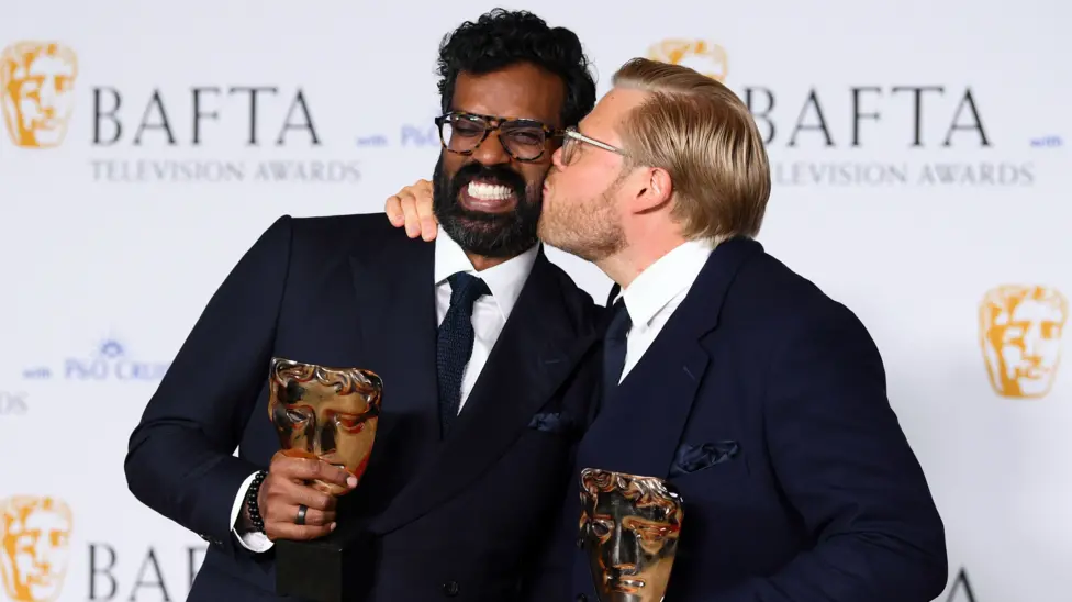 Romesh Ranganathan and Rob Beckett posing at the 2024 Baftas. Rob is kissing Romesh on the cheek