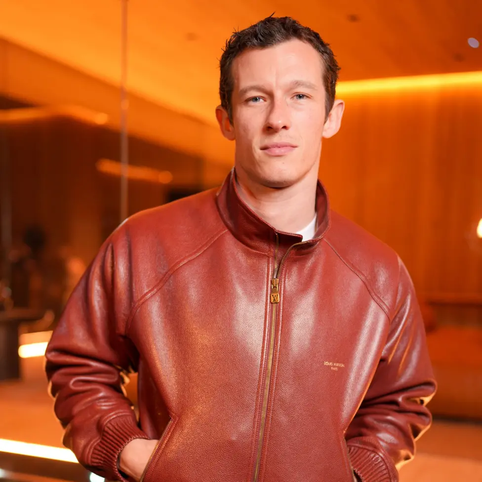 Callum Turner pouts as he looks at the camera posing for a photo wearing a brown leather jacket in a club like setting, in Paris in January.