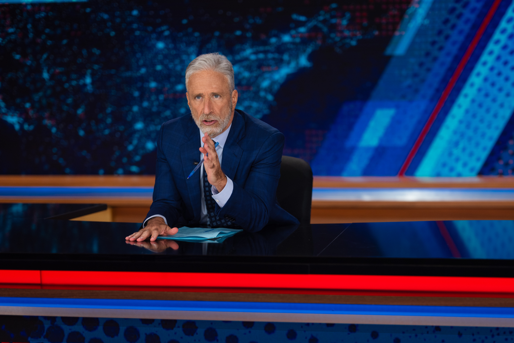 Jon Stewart seated behind a news desk delivering a late-night comedy monologue