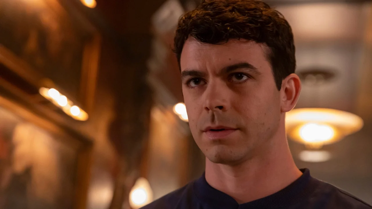 Close-up of a man with short curly dark hair and a navy top, looking intently to the side in a warmly lit hallway.