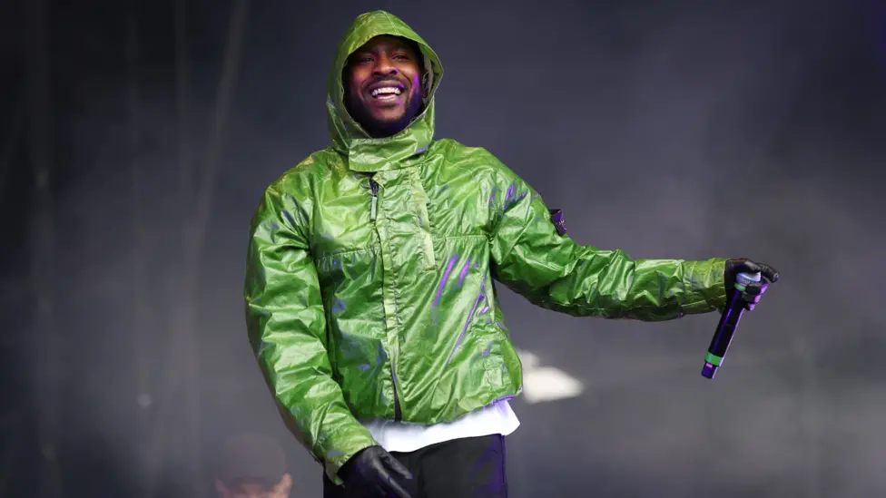 Skepta performs on stage wearing a green jacket, black gloves and black trousers.