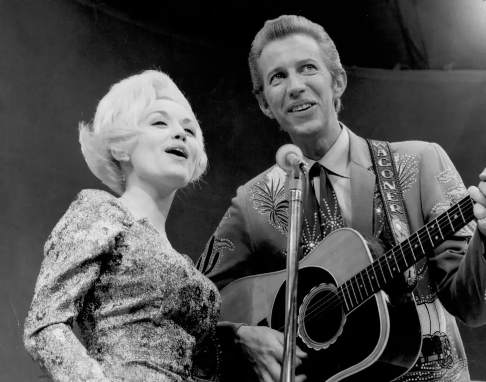 Black and White image of Dolly and Porter singing into a shared microphone, and he is playing a guitar.