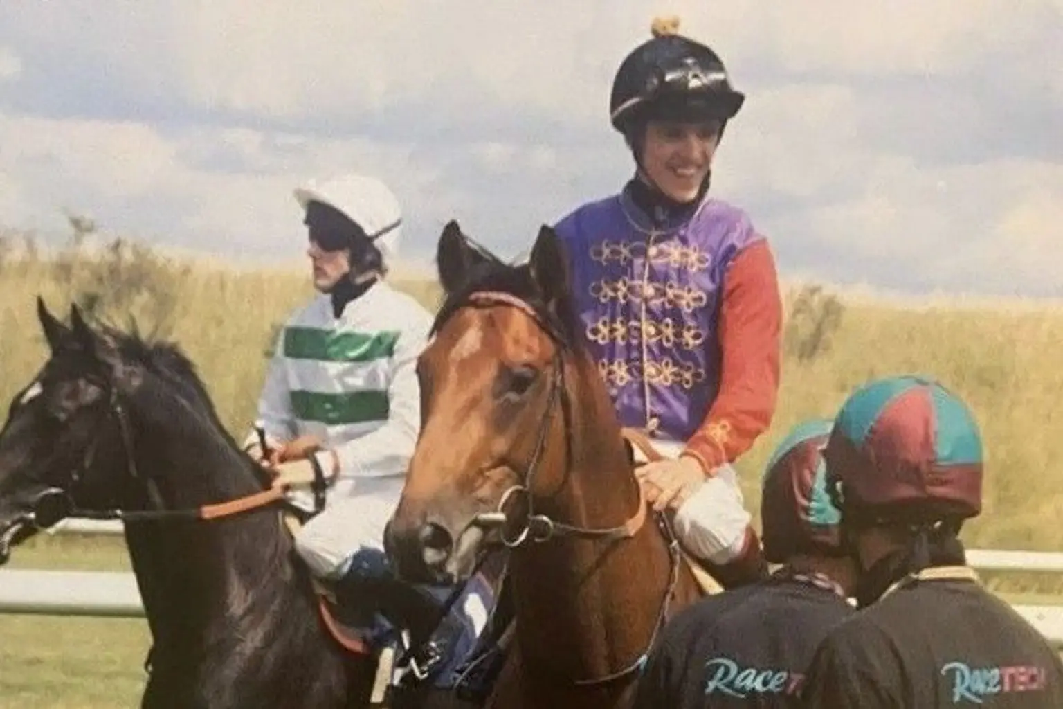 JJ Hamblett is riding a brown horse and is wearing purple and red jockey colours and a black helmet. He is talking to two people stood on his left.