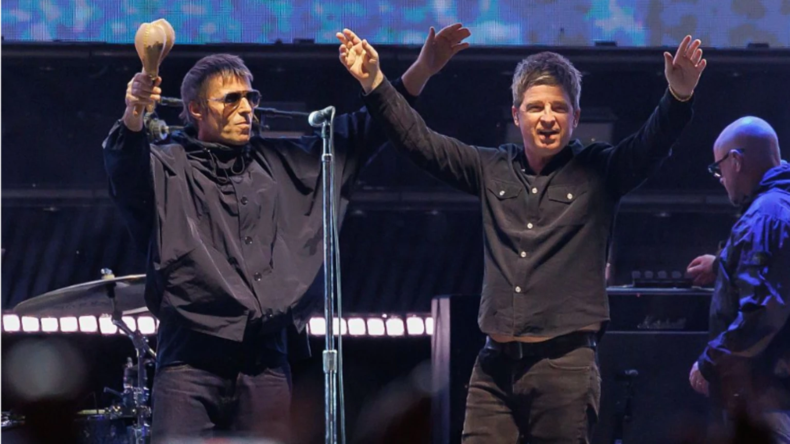 Oasis on stage in 2025. Both Liam and Noel are wearing black shirts and raising their arms into the air to greet fans. Liam is holding a pair of maracas in his right hand.