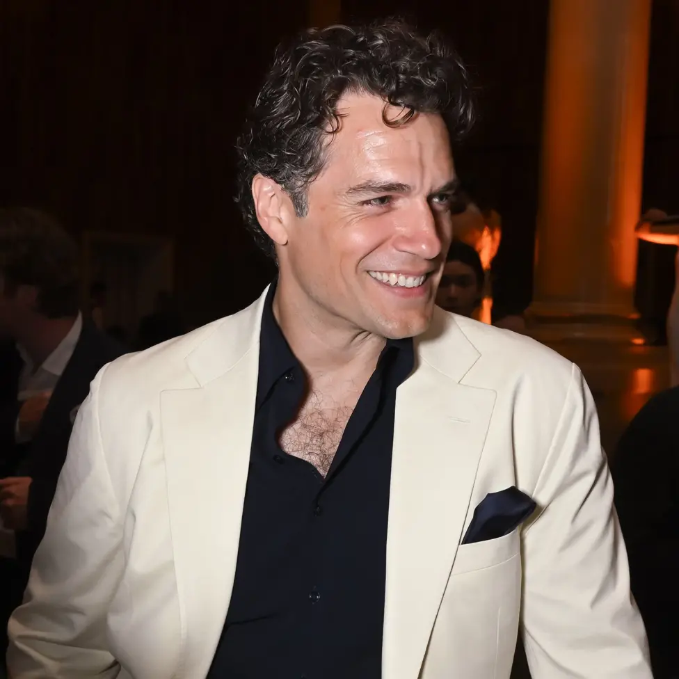 Henry Cavill smiles looking to right of frame while wearing a white suit jacket and black shirt, in London in June.