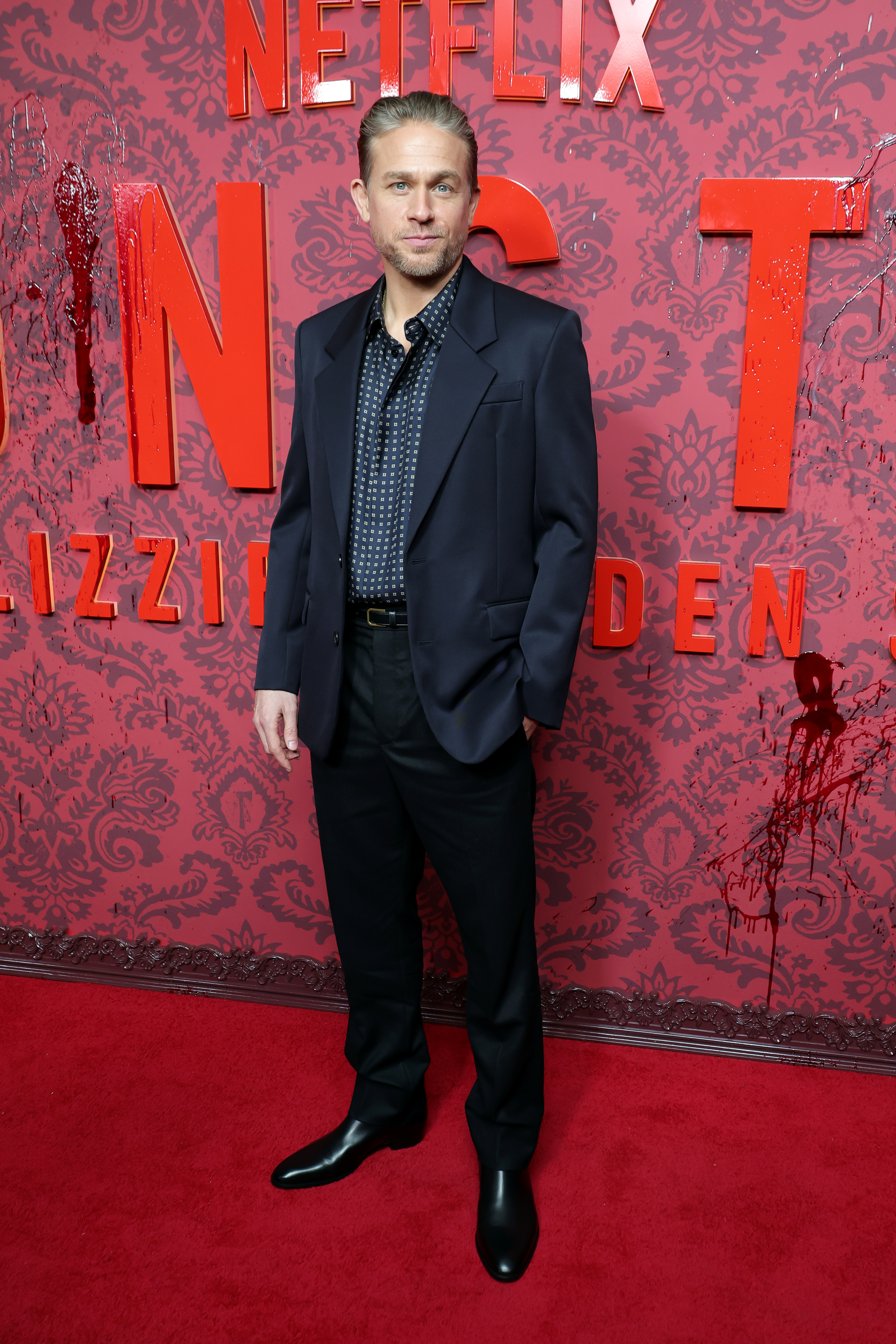 Charlie Hunnam poses on a red carpet in front of a red patterned backdrop for Netflix