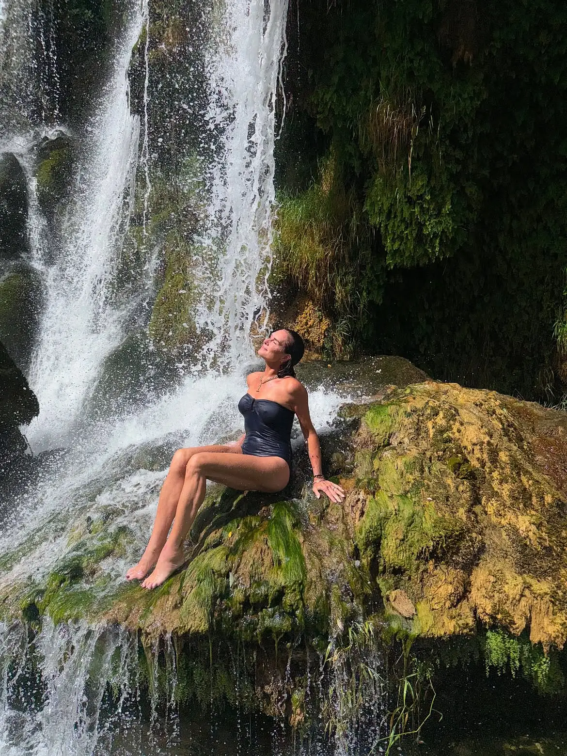 Brooke Shields posing in a navy strapless swimsuit under a waterfall in Croatia.