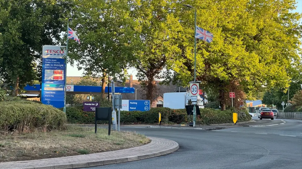 A Tesco filling station - mostly obscured by tall trees, but the prices sign is in the foreground just off a roundabout. There is a union jack flag flying from a lamp-post, above a 30mph speed limit ending sign.
