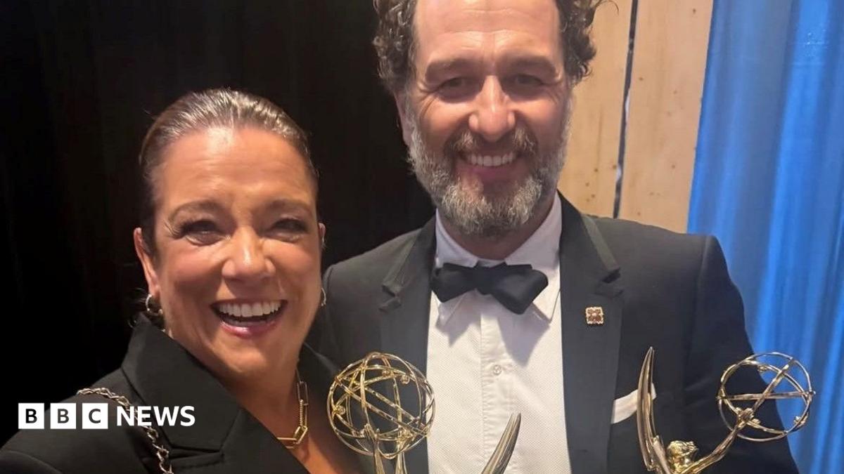 Rachel wears a black suit and gold necklace, her hair tied back. Matthew wears a black suit and white shirt. They are smiling and holding two Emmy awards