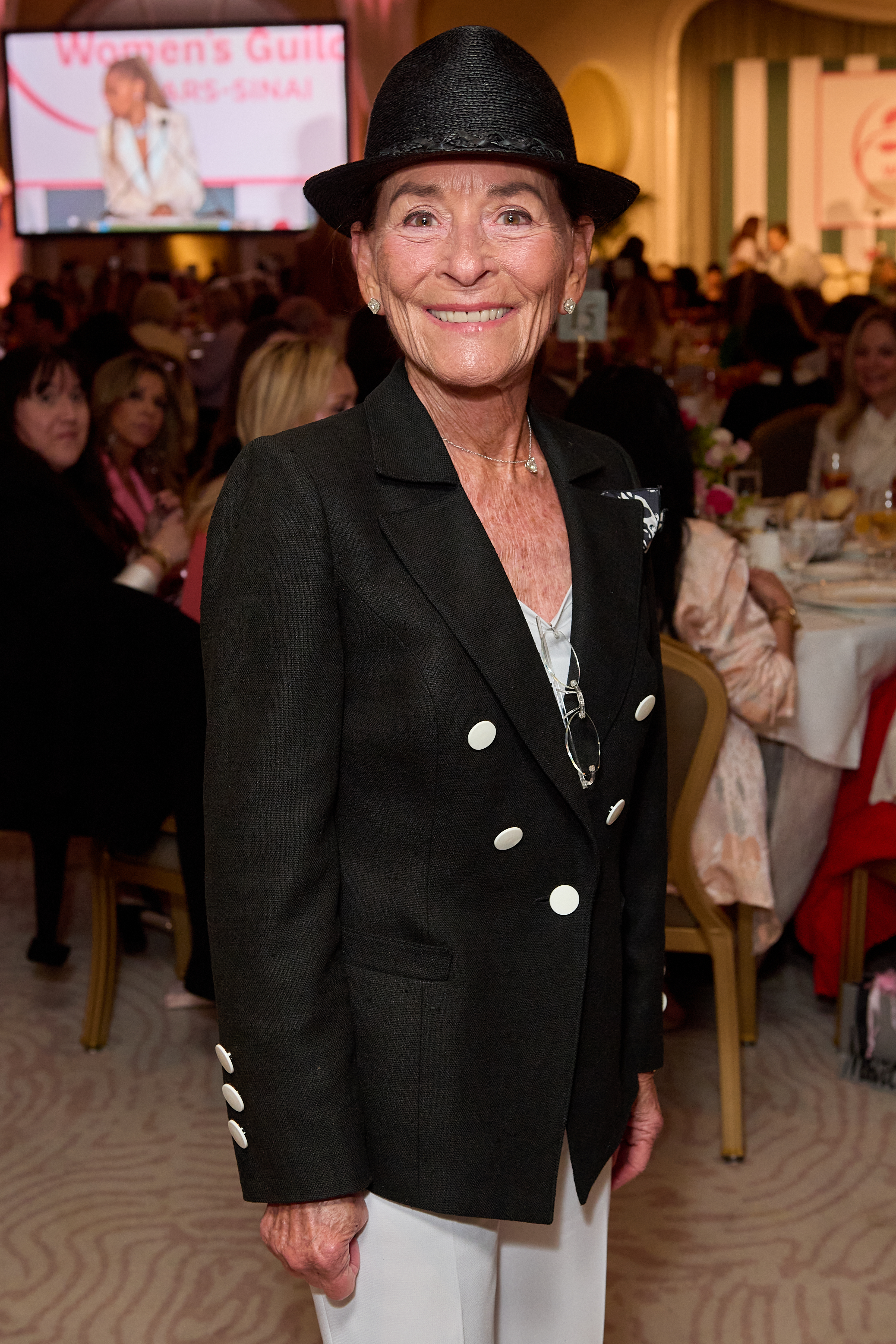 Judge Judy Sheindlin smiles wearing a black blazer, white pants, and a fedora.