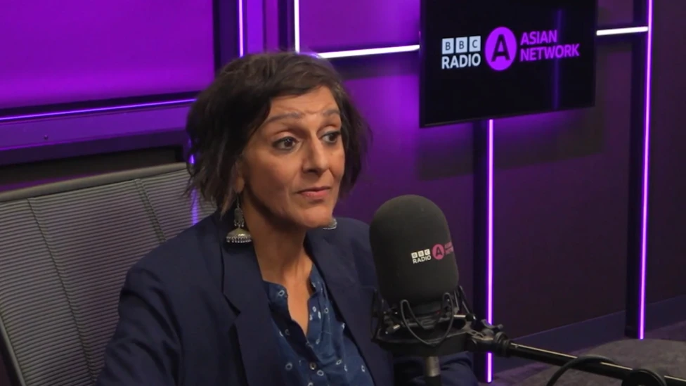 Dame Meera Syal a woman with chin-length brown hair and long silver earrings is wearing a navy blue blazer over a navy blue shirt with small white flowers on. She is sat behind the desk in a radio studio talking into a m