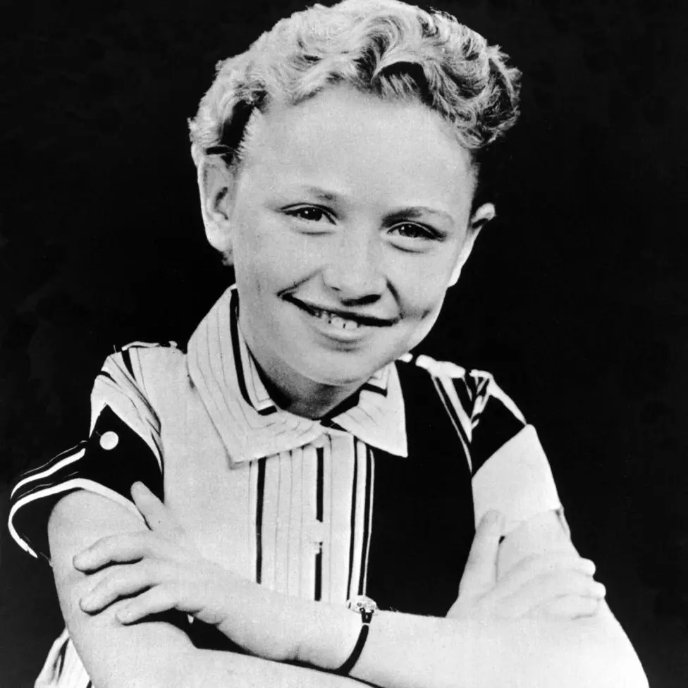 A black and white photo of Dolly as a child. She is sitting at a desk smiling and her hair is very short and curly. She wears a conservative black and white striped short sleeved shirt buttoned up to her neck.