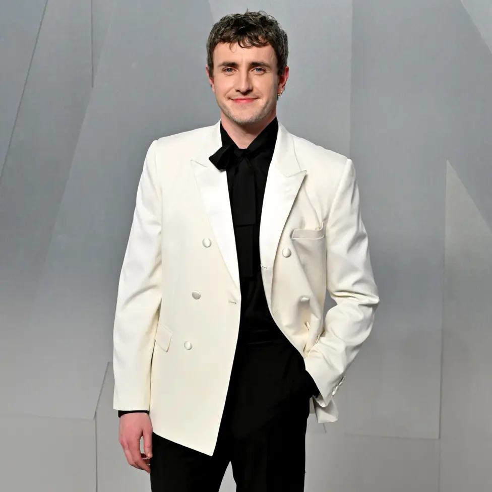 Paul Mescal smiles while posing for a photo wearing a white tuxedo with black shirt and trousers against a grey background, in LA in March.