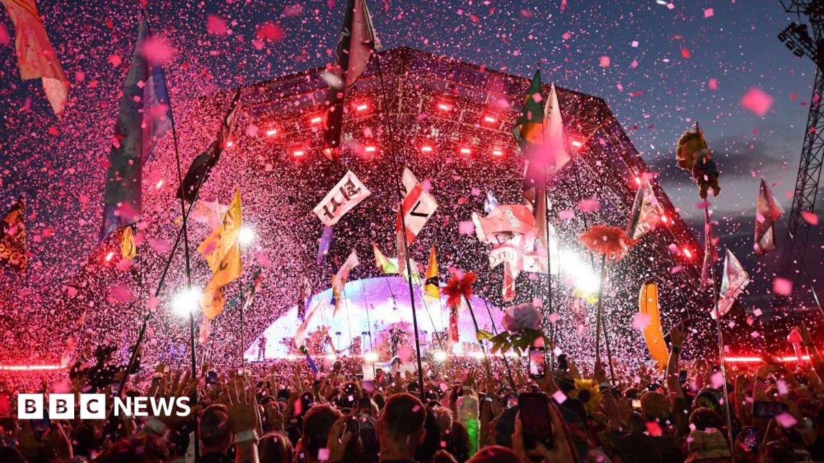 The Pyramid stage at Glastonbury festival is pictured lit up with a pink hue at night. There are thousands of pieces of confetti flying through the air as people in the crowd wave their arms and hold up tall flags.