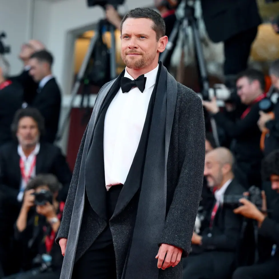 Jack O'Connell looks towards camera while posing on the red carpet wearing a black tie outfit in Italy in September.