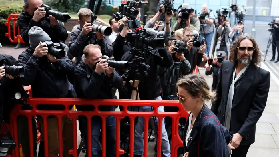 Russell and Laura Brand holding hands as they walk past a group of photographers behind a red barrier outside court