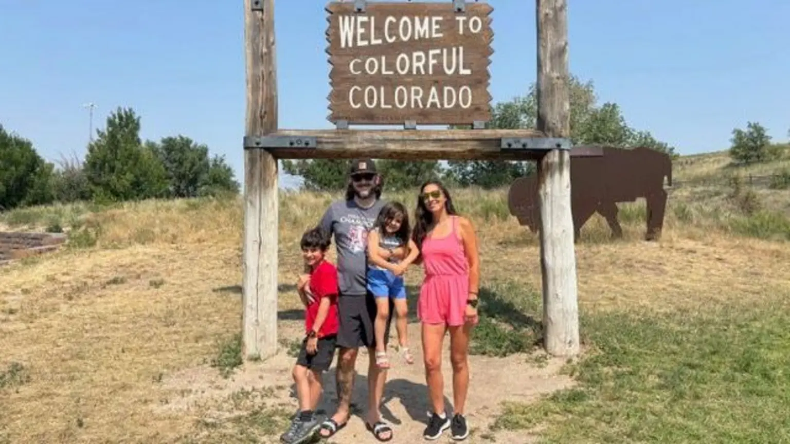 Colorado mother Ciara Fernandez Faber, wearing a short pink romper, stands with her husband, son and daughter under a sign that reads 'Welcome to Colourful Colorado' - with grass, shrubbery and a bison art piec