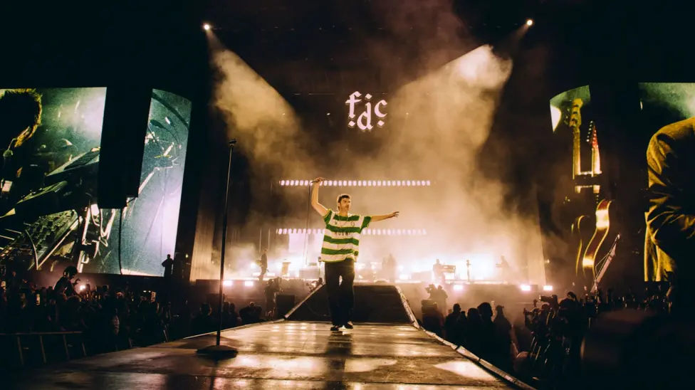 A landscape shot of Fontaines D.C. performing at Reading Festival. Grian Chatten is centre stage and seen on two large screens to each side of the stage, performing whilst wearing a Cwmbran Celtic FC away shirt.