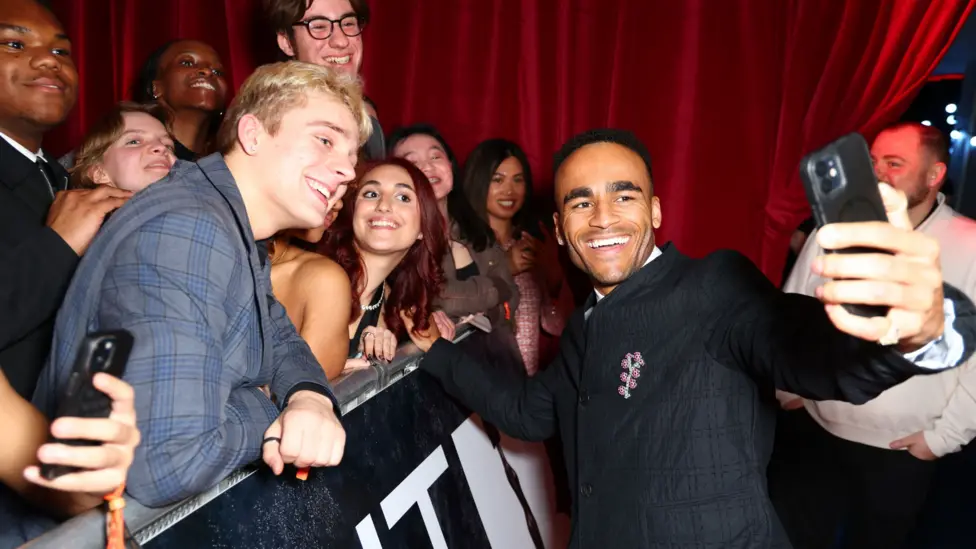 Munya Chawawa smiling, holding a phone and taking a selfie with a group of fans on the other side of a barrier on the red carpet at a Brit Awards event in 2025