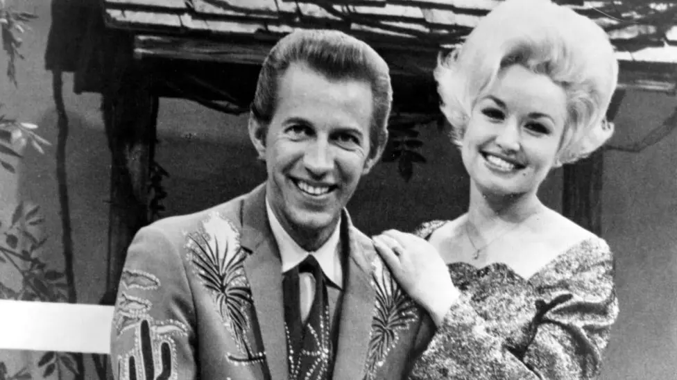 Wagner sits on a stool in a suit with emblems of cacti and plants in this black and white photogragh. He is posing with his guitar. Dolly stands behind him leaning on his shoulder. Her hair is short but still standing on