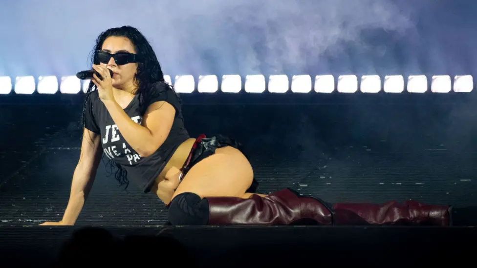 Charli XCX performing on stage. She is holding a microphone and wearing a black t-shirt, a black short skirt and red boots.