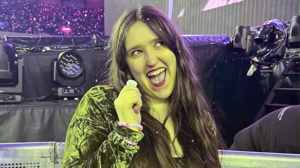 Georgia McAdam smiles while holding a guitar pick. She has long dark hair and wears a velvet jacket. She's pictured next to a stage, lit by a green light.