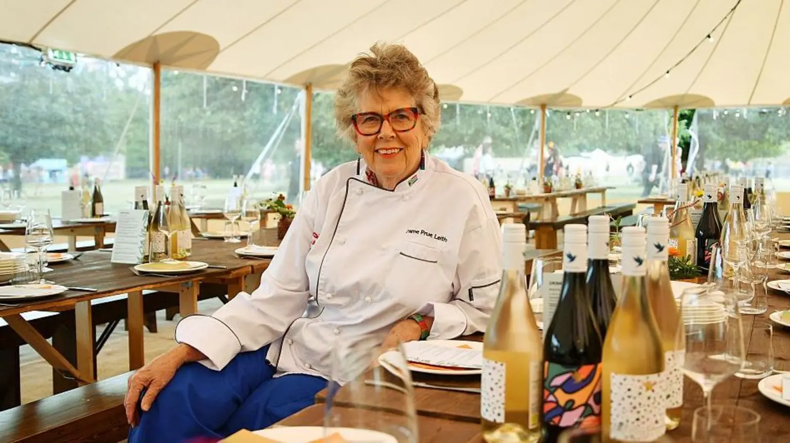 Dame Prue Leith, who is wearing a white chef's shirt and blue trousers, sits down at a long table in the dining area of a festival. She is smiling and has short hair and glasses. There are bottles of wine, wine glas