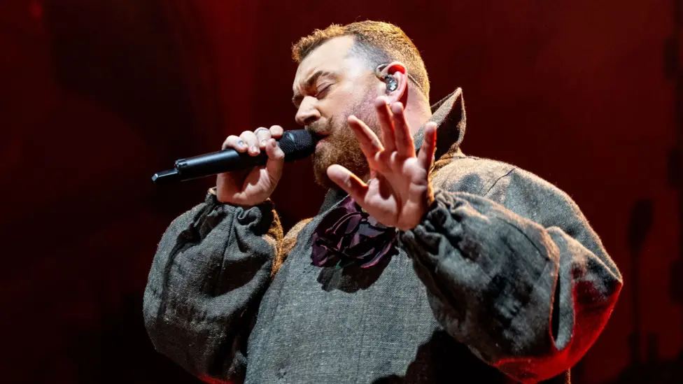 Sam Smith sings into a microphone while performing at the Albert Hall. They wear a grey coat with a purple flower pinned on the front.