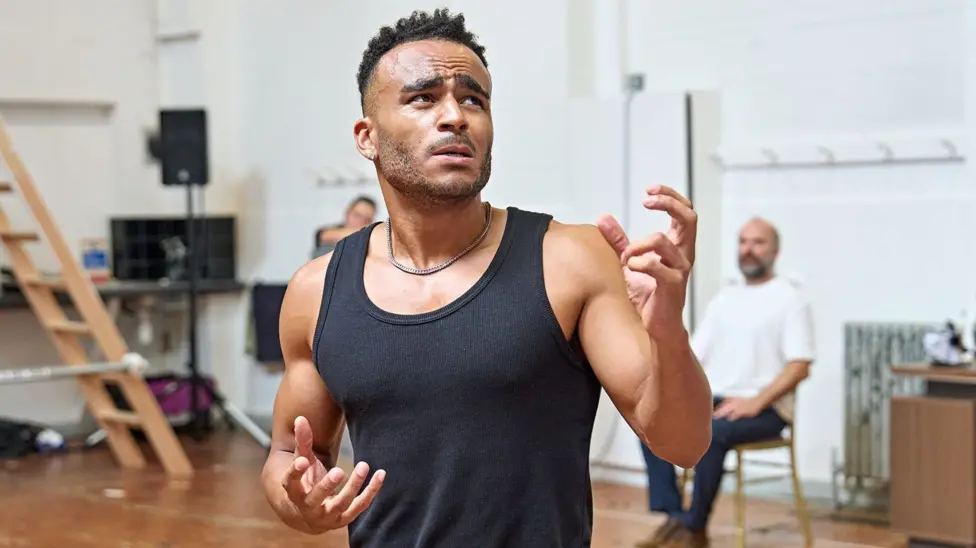 Munya Chawawa in a black vest with both arms gesturing and his face with a pained expression, in a rehearsal room