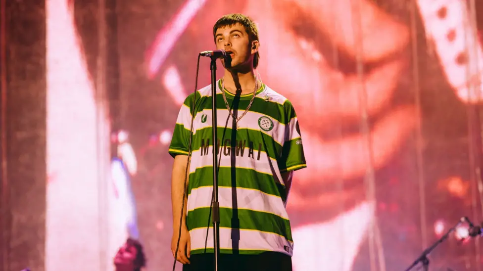 Grian Chatten performing at Reading Festival wearing a Cwmbran Celtic FC away shirt. The football shirt is green and white.