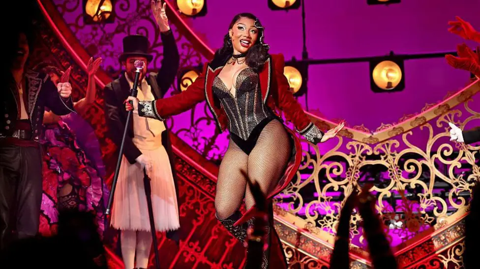 Megan is wearing a red cropped jacket, a black sparkly leotard with silver stripes. She is wearing fishnet tights and black knee high boots. She is holding a black cane and smiling a the crowd. She is on stage, behind he