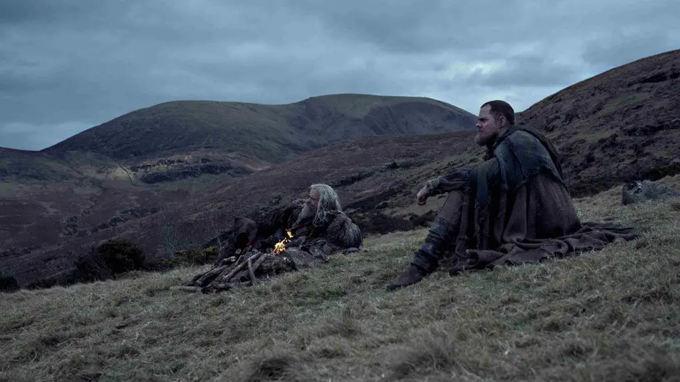 Two men in brown medieval sackcloth clothing, looking off into the distance. They are lying/sitting on grass, beside a fire. There are green and brown hills and fields behind them.